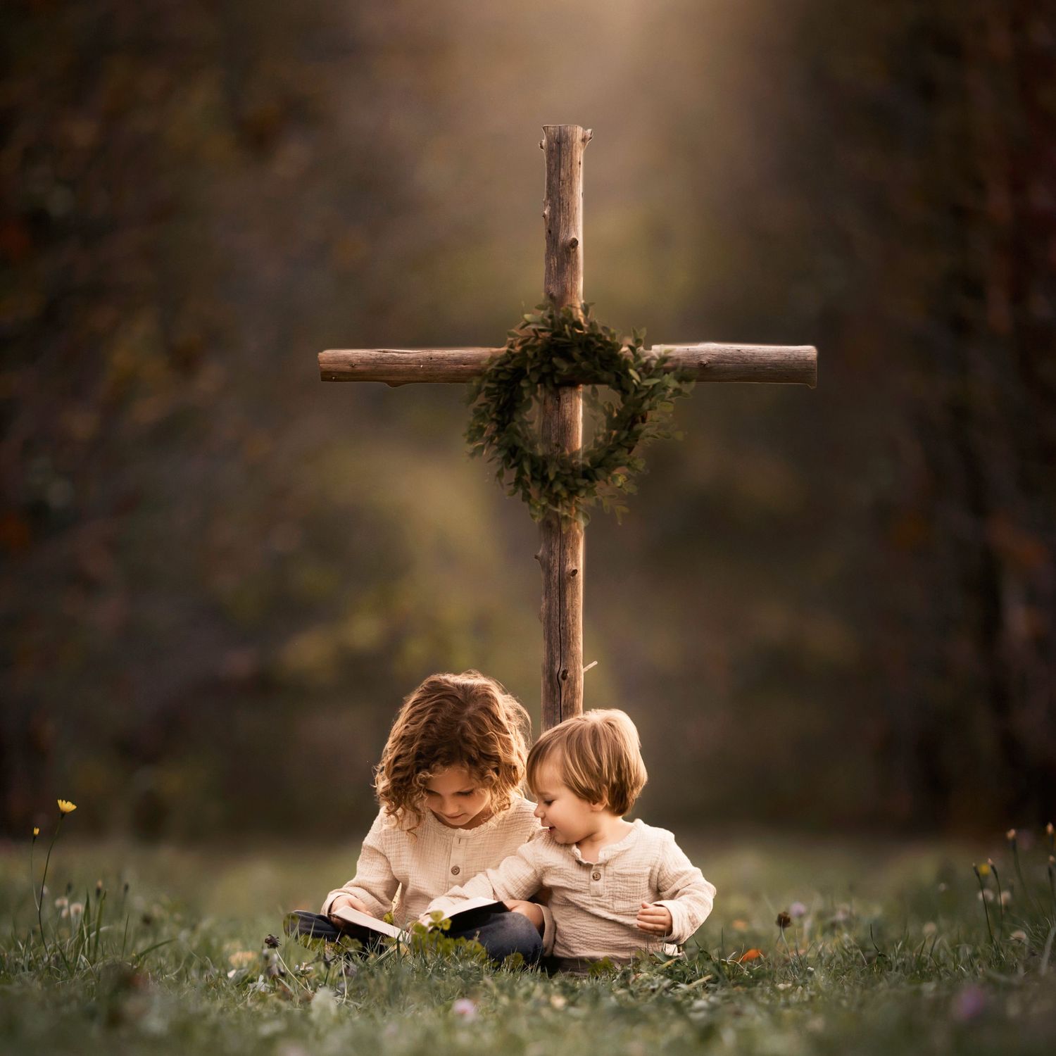Two children reading together beneath a wooden cross adorned with greenery.