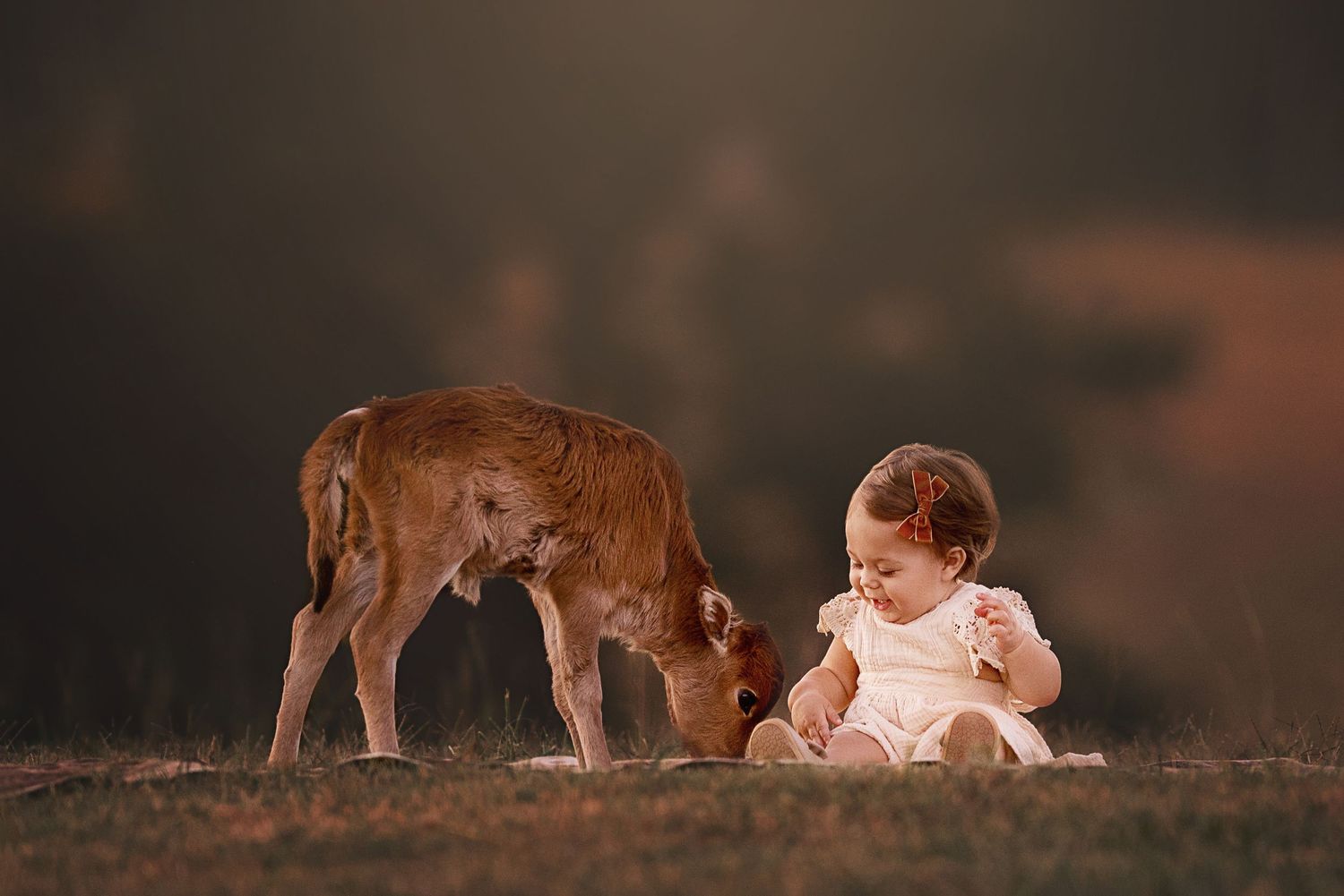 A young deer and a child in a white dress sit together in a meadow during golden hour.