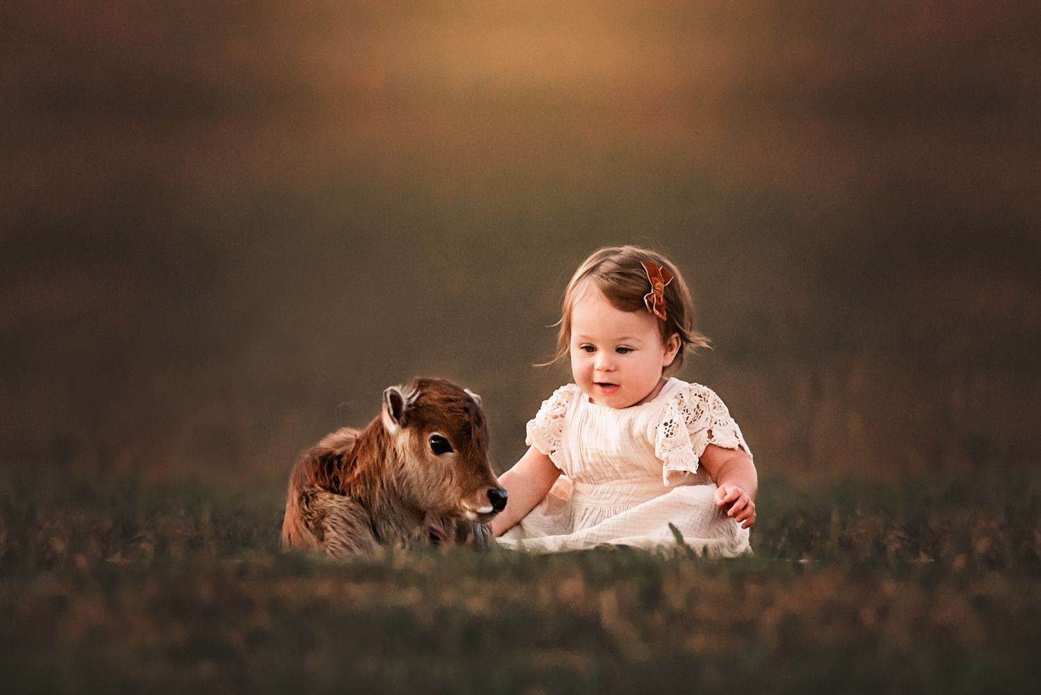 A small goat and a child in white lace dress sit together in a grassy field at dusk.
