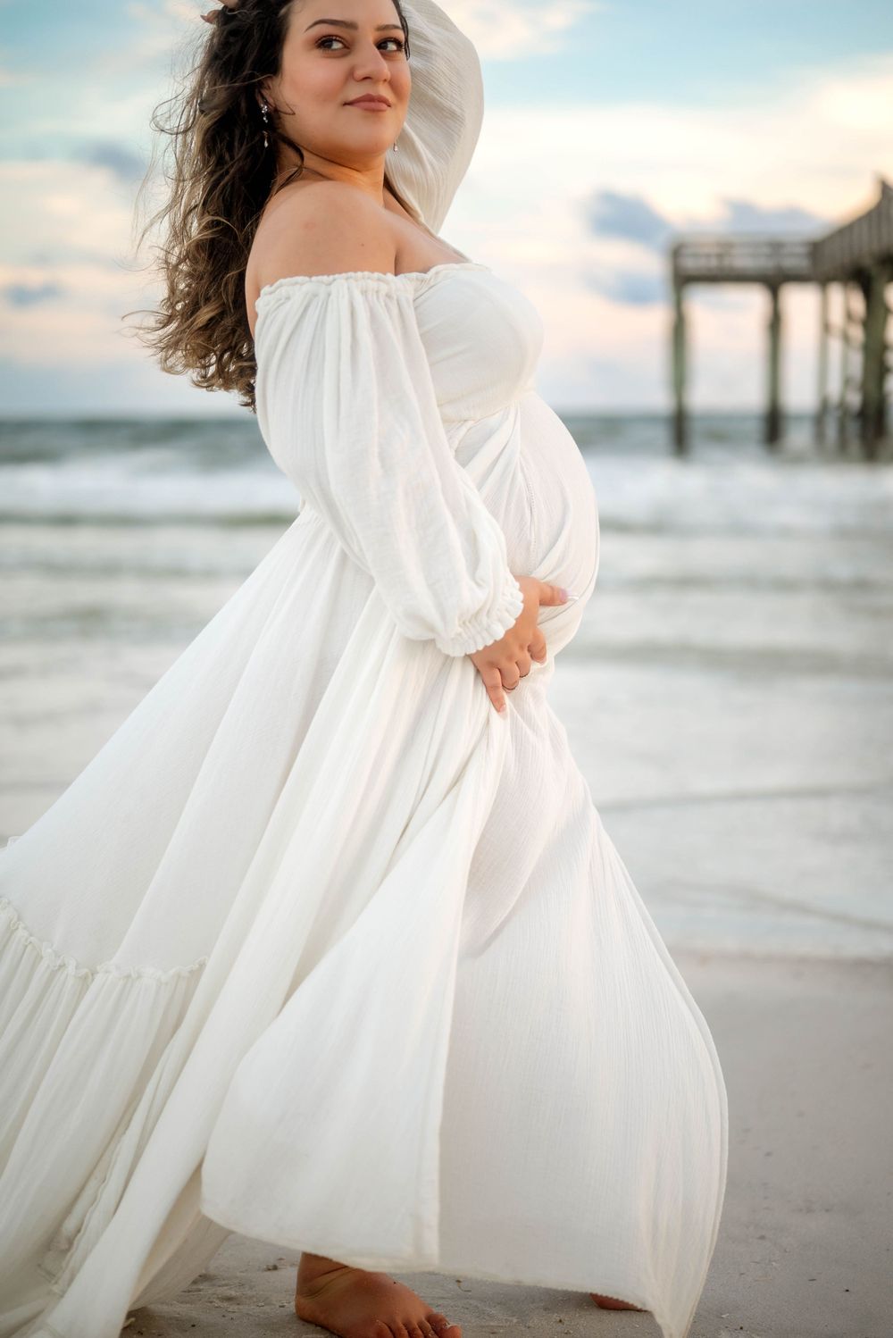 Flowing white dress catches ocean breeze during beach maternity photoshoot.