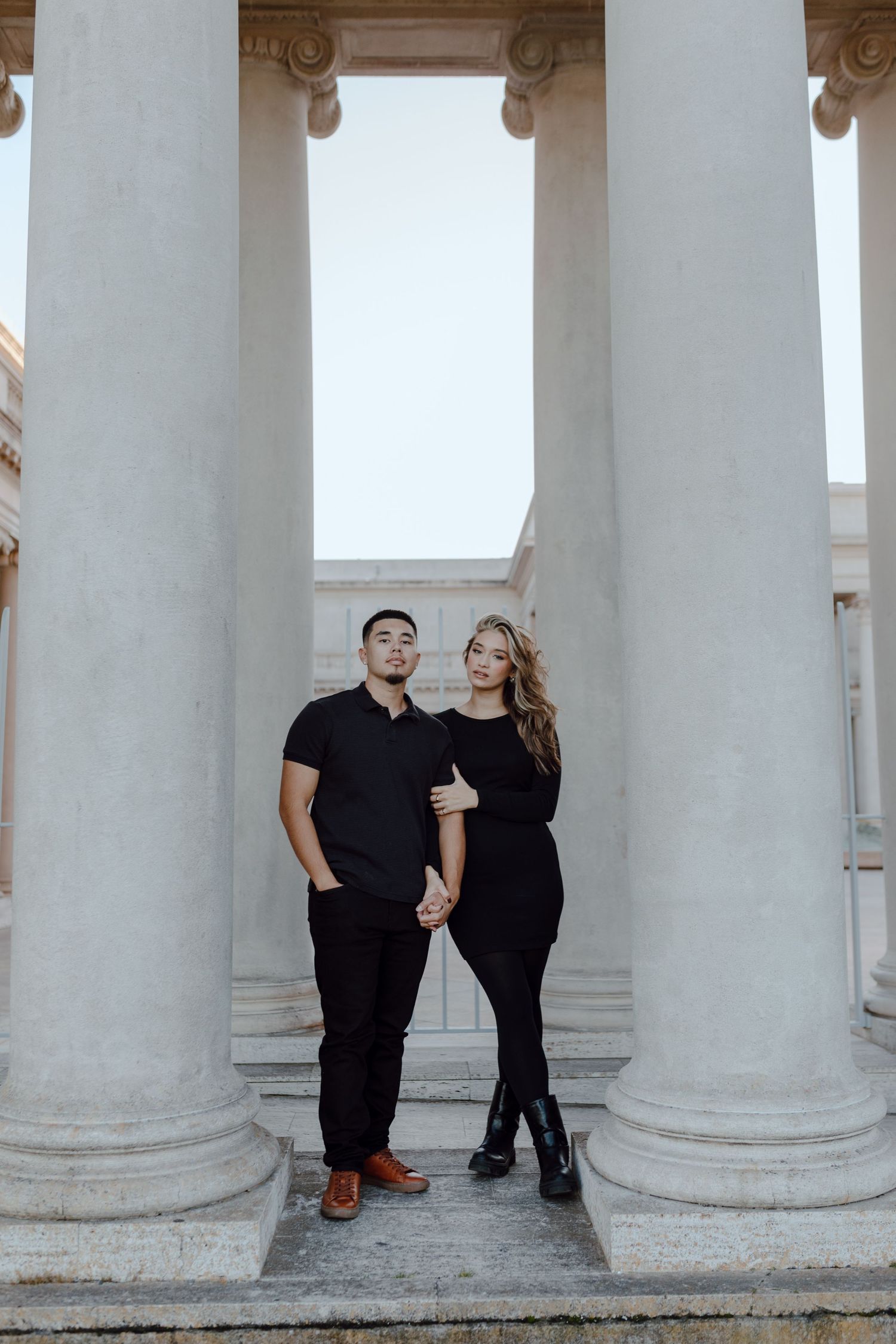 A couple poses between classical white columns in matching black outfits.