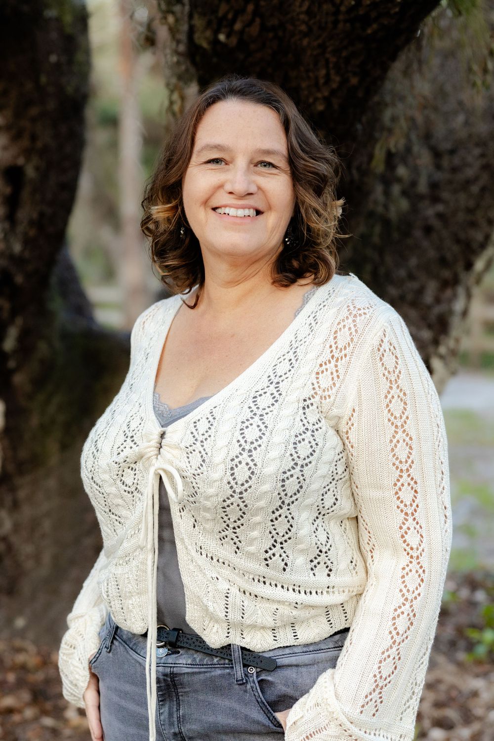 Person wearing cream crochet cardigan and jeans poses against tree outdoors.