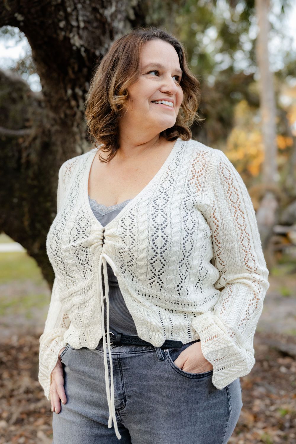 Person in cream lace cardigan and jeans standing by tree trunk outdoors.