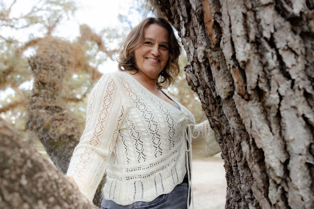 Person in cream knit cardigan poses beside large tree trunk outdoors.