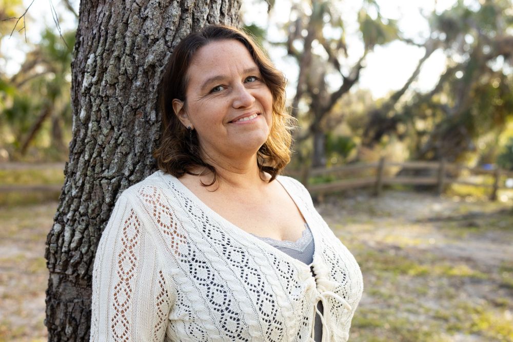 Portrait of someone in white knit cardigan standing against a tree trunk.