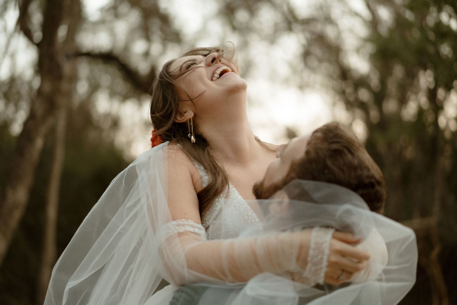 A joyful wedding moment captured in natural light with flowing white veil against a forest backdrop.