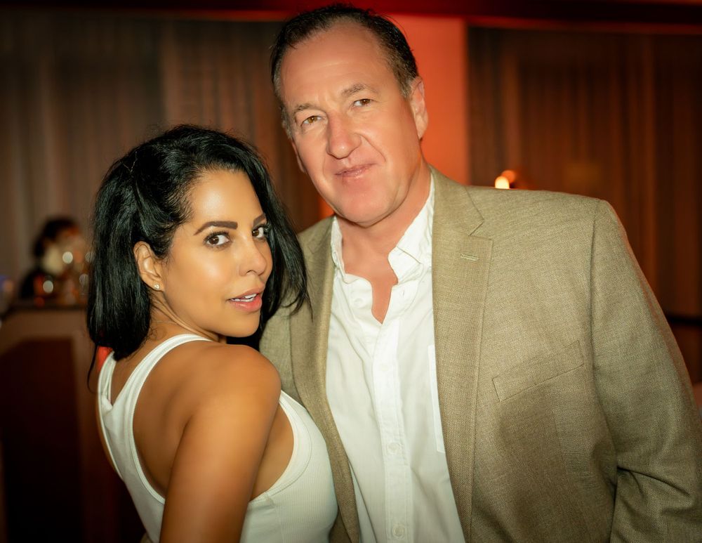 A person in a white tank top poses with someone wearing a beige suit jacket at an indoor event with warm lighting.