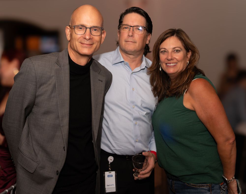 Three people pose together at an indoor social event, with two wearing business attire and one in casual green attire.