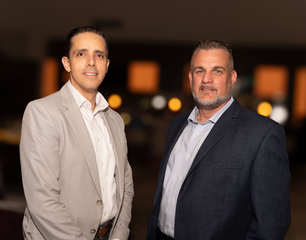 Two business professionals in formal attire stand together at an evening event with warm lighting in the background.