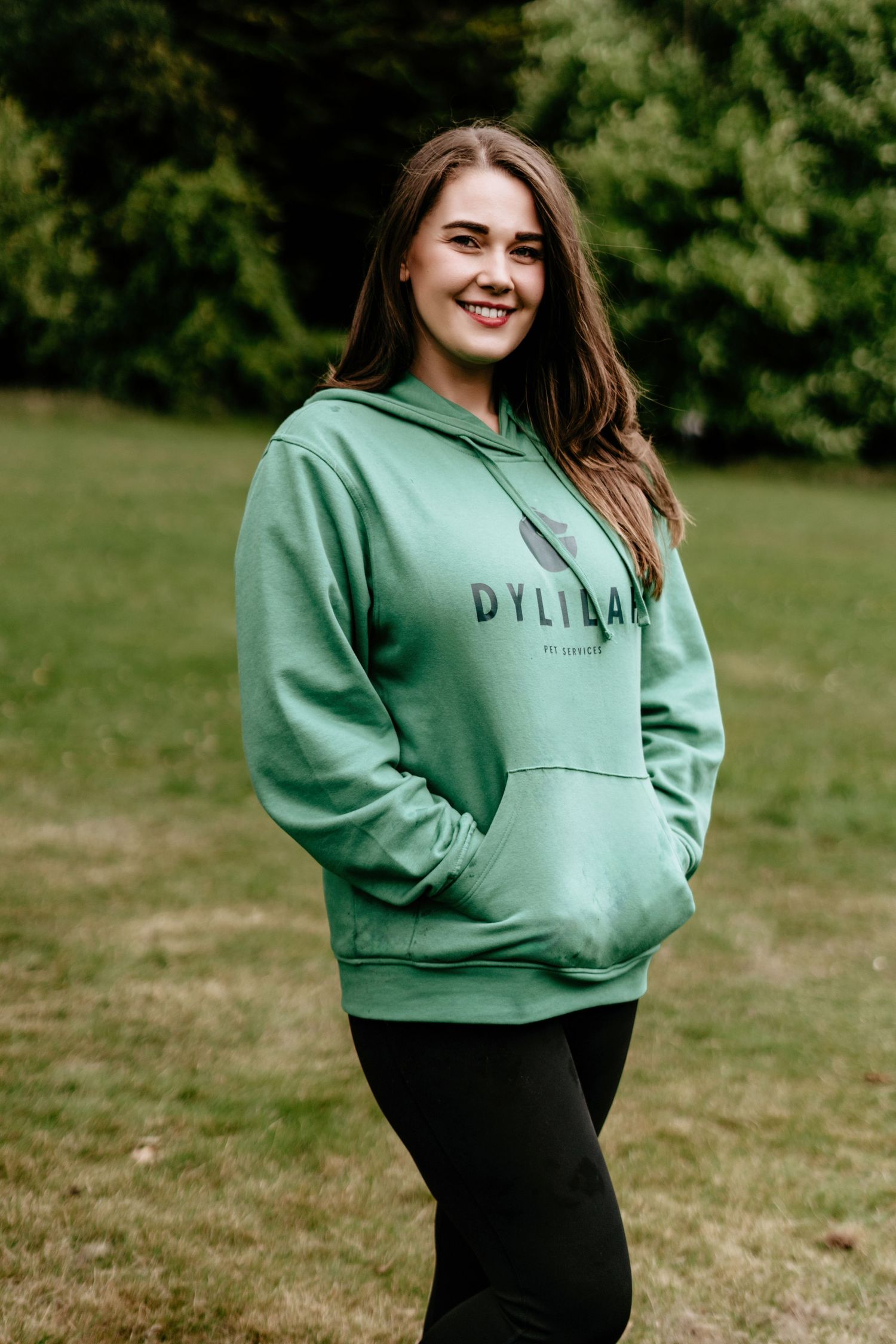 Professional pet service worker wearing mint green hoodie standing in a grassy outdoor setting.