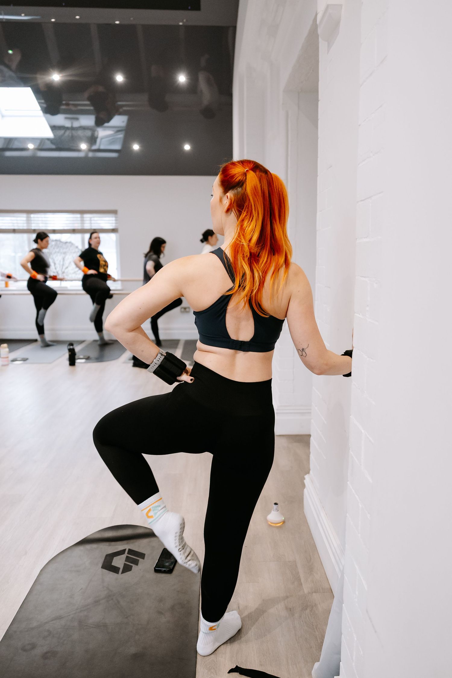 Person with bright orange hair wearing black workout attire exercises in a modern dance studio with mirrors.