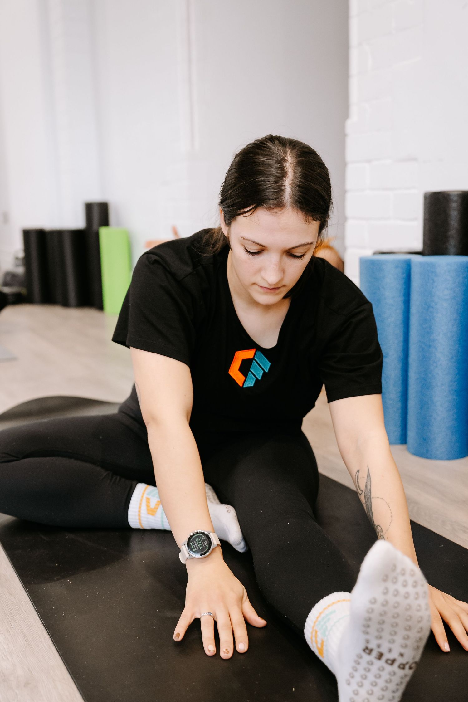 A person in a black t-shirt with a logo stretches on a mat at a fitness studio with foam rollers in the background.