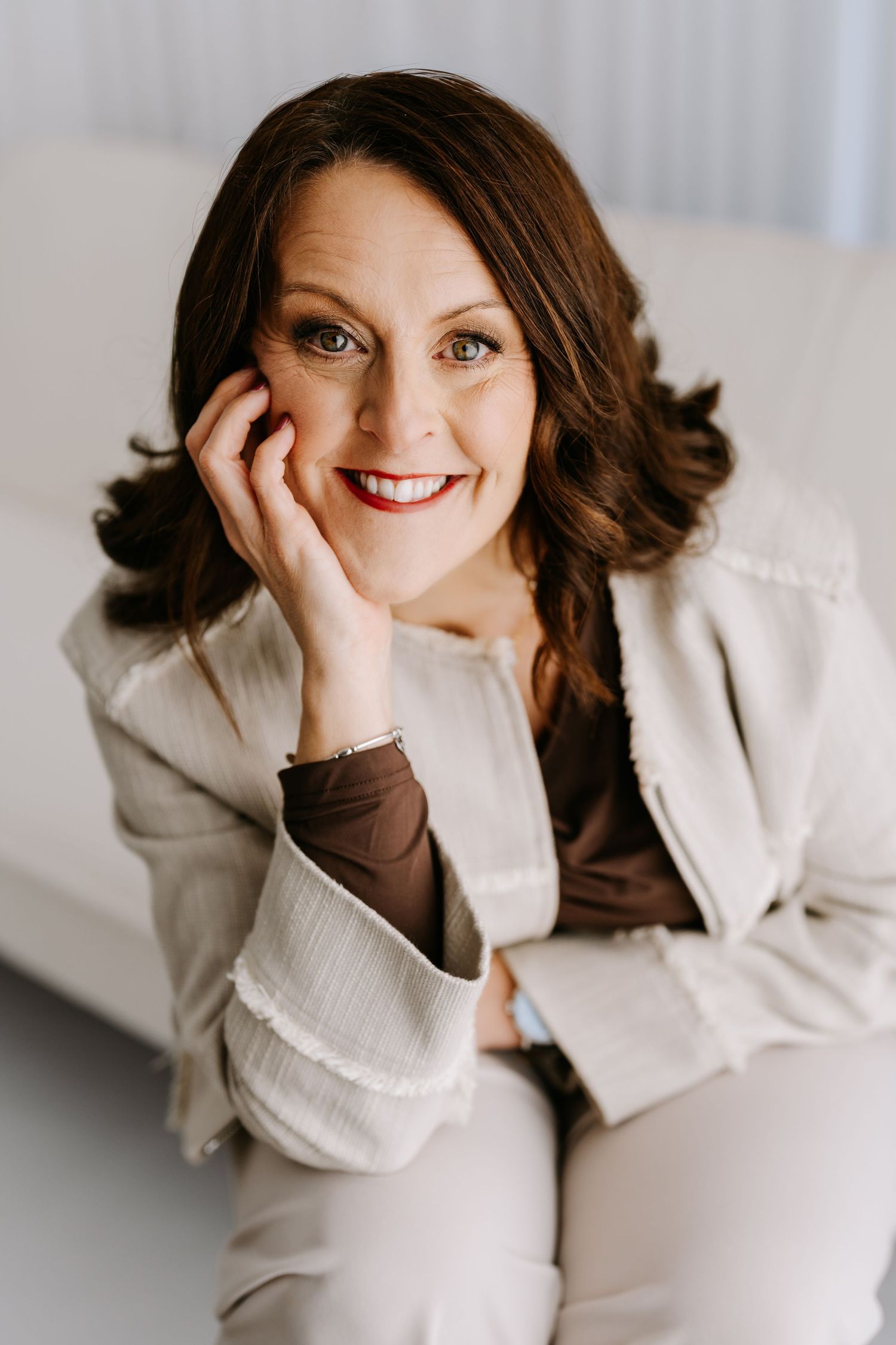Professional portrait of someone in a cream colored blazer and brown shirt smiling warmly while resting their face on their hand.