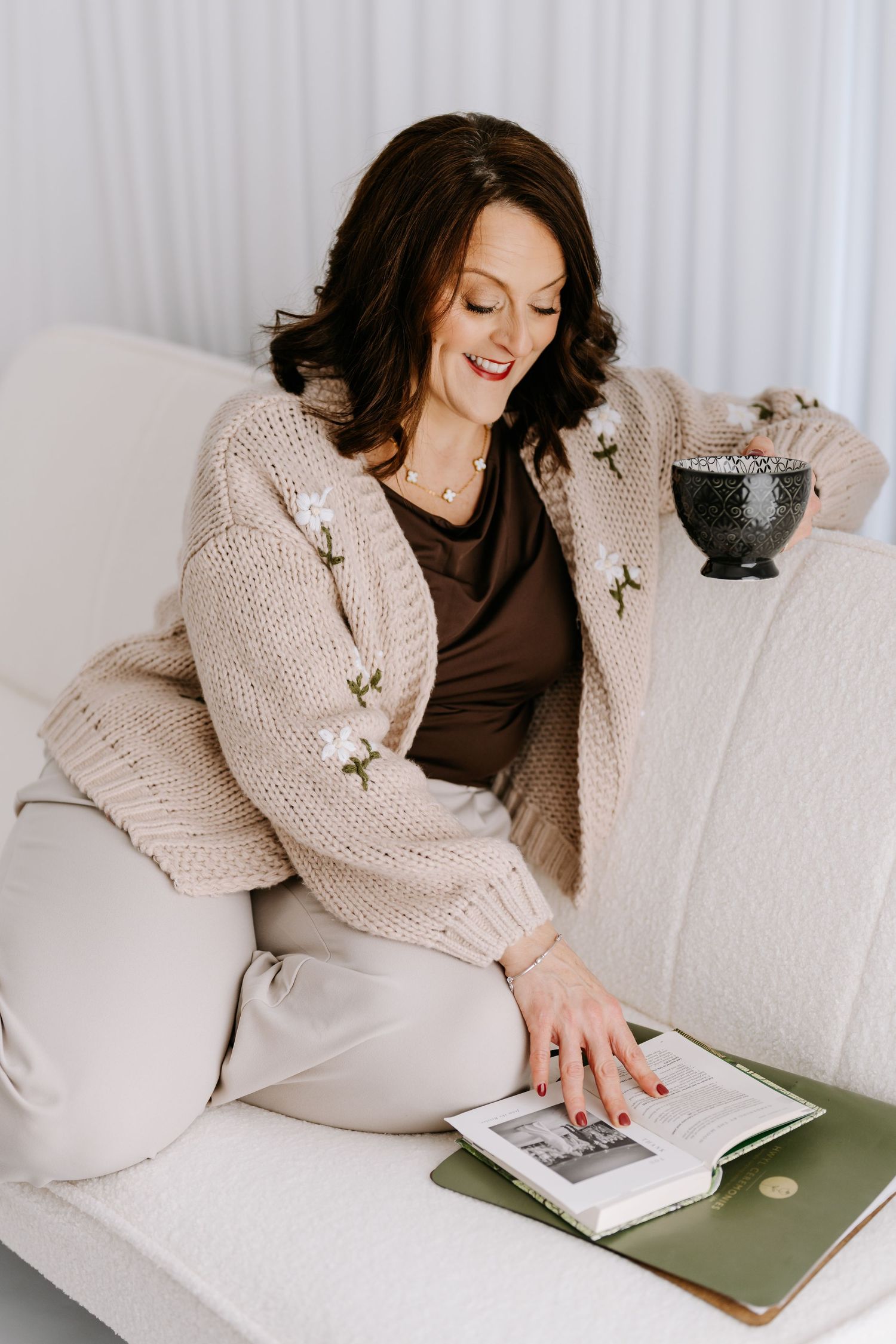 Person relaxing on white couch wearing cozy knit cardigan while reading and holding coffee mug.