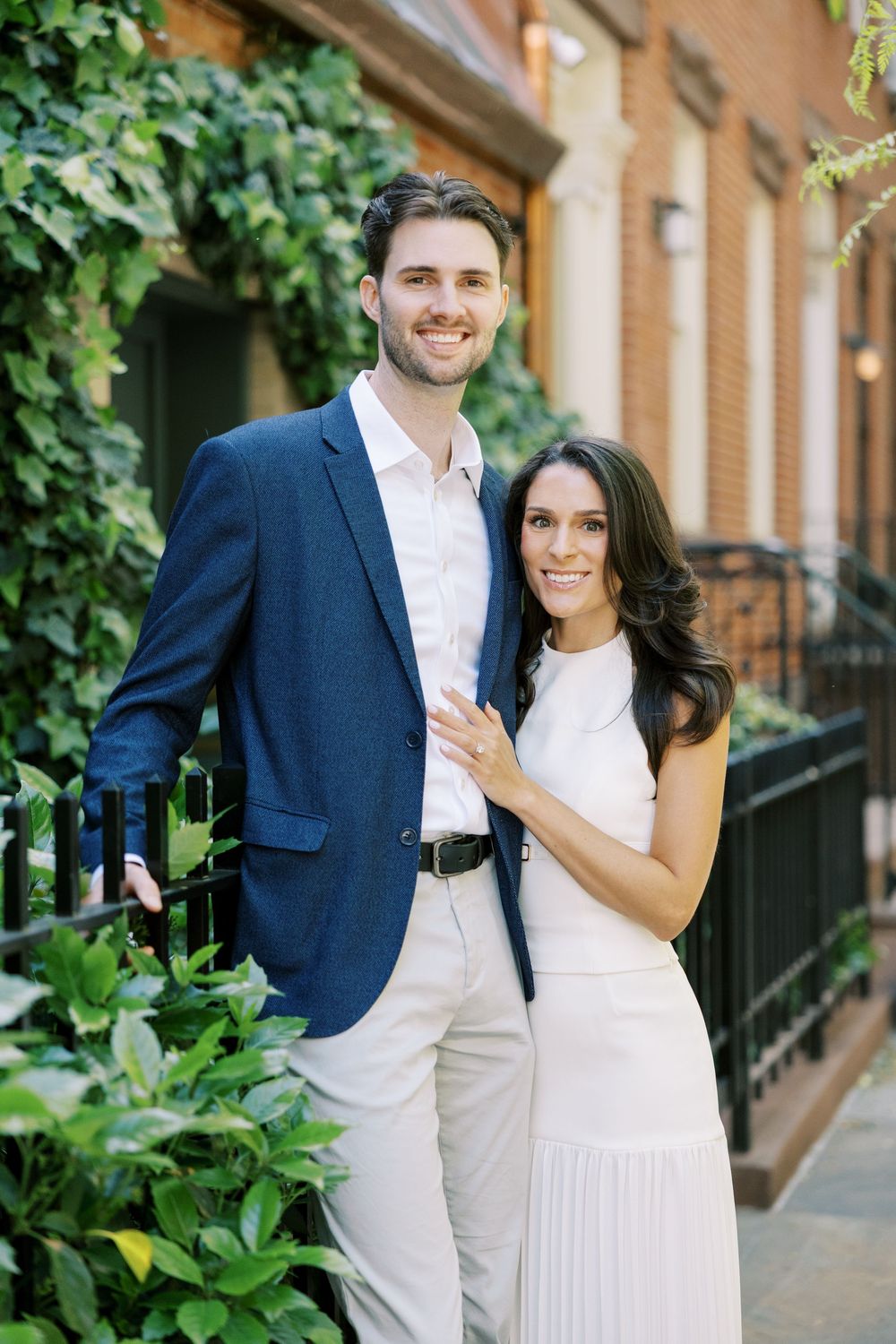 A couple poses elegantly against a brick brownstone with ivy, wearing a navy blazer and white pleated dress in New York City.