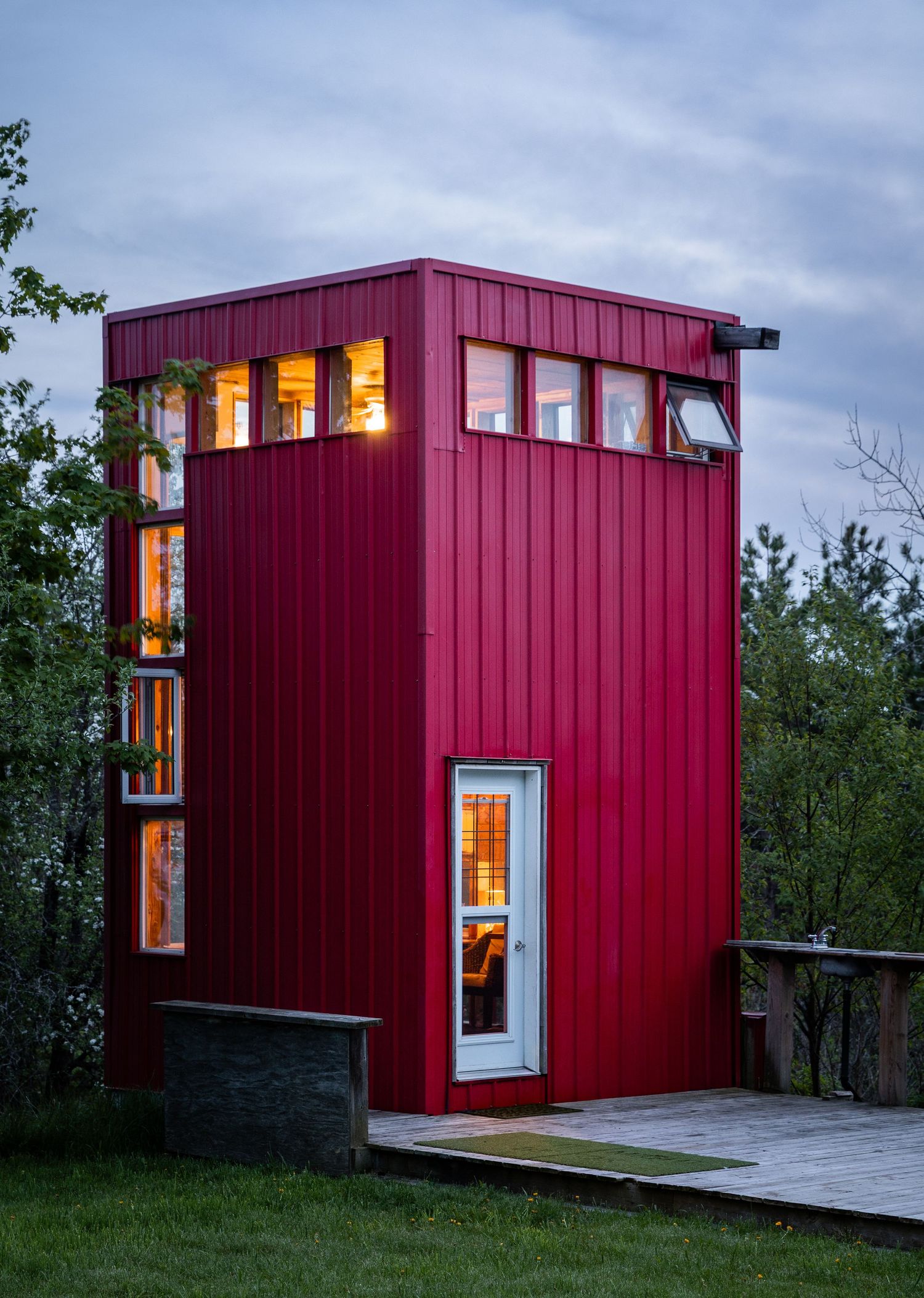 A tall red metal-clad tiny house glows warmly at dusk, surrounded by green trees and a wooden deck.