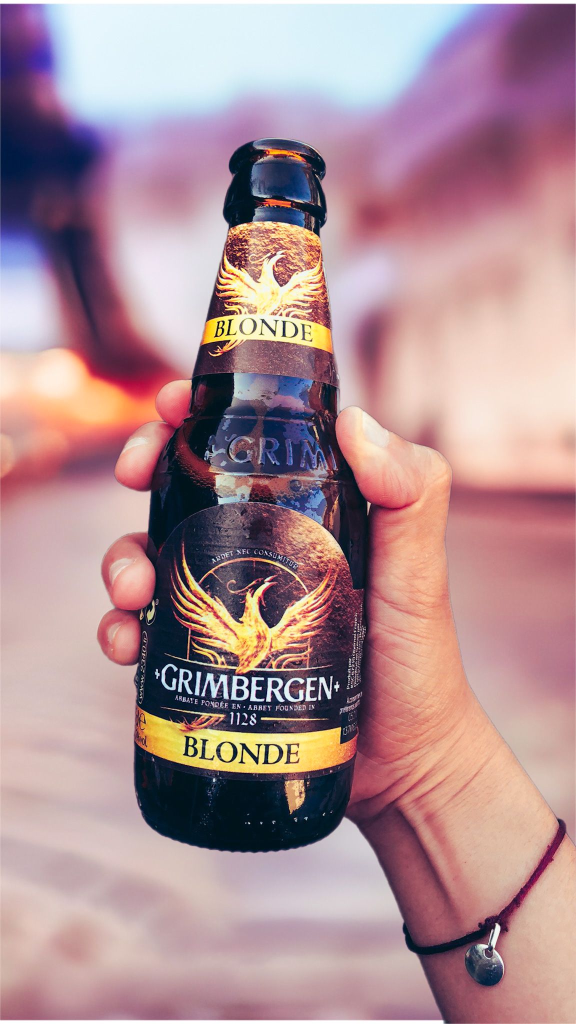 Hand holding a Grimbergen Blonde beer bottle with golden phoenix logo against a blurred outdoor background in Paris.