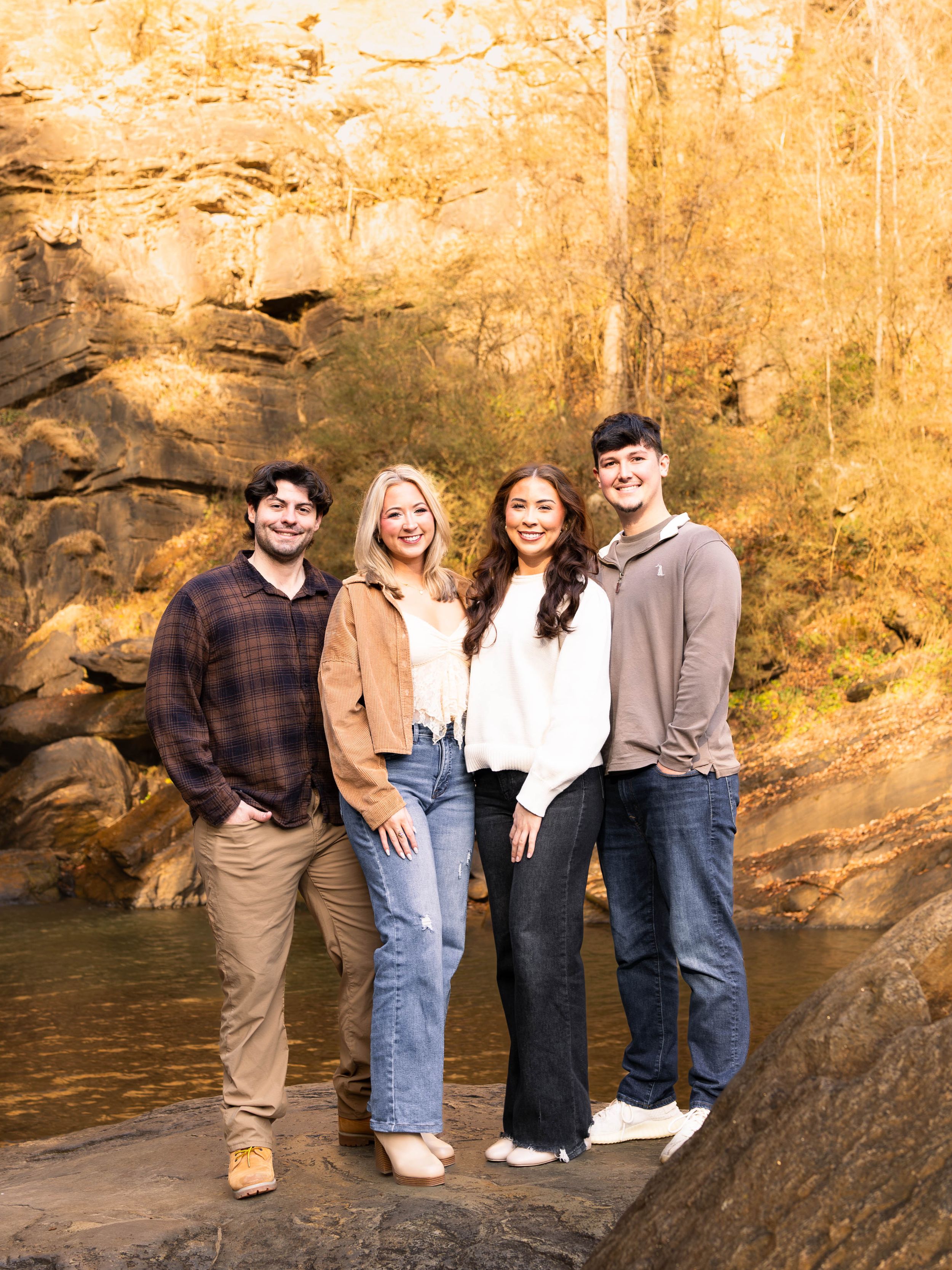 Four people sitting casually on large rocks by a river during golden hour in an autumn outdoor setting.