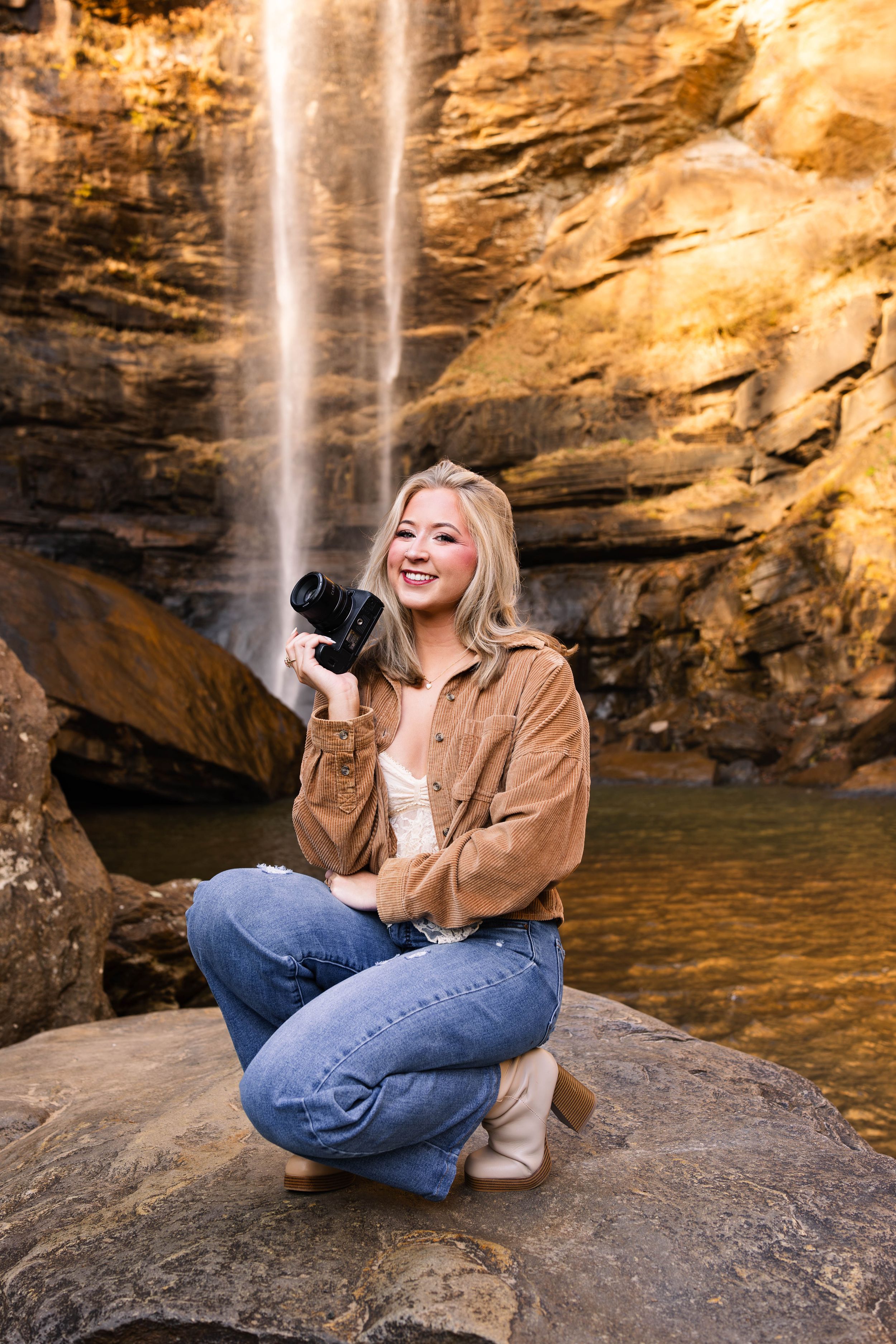 A person sits on a rocky ledge near a cascading waterfall while holding a coffee mug and enjoying the natural scenery.