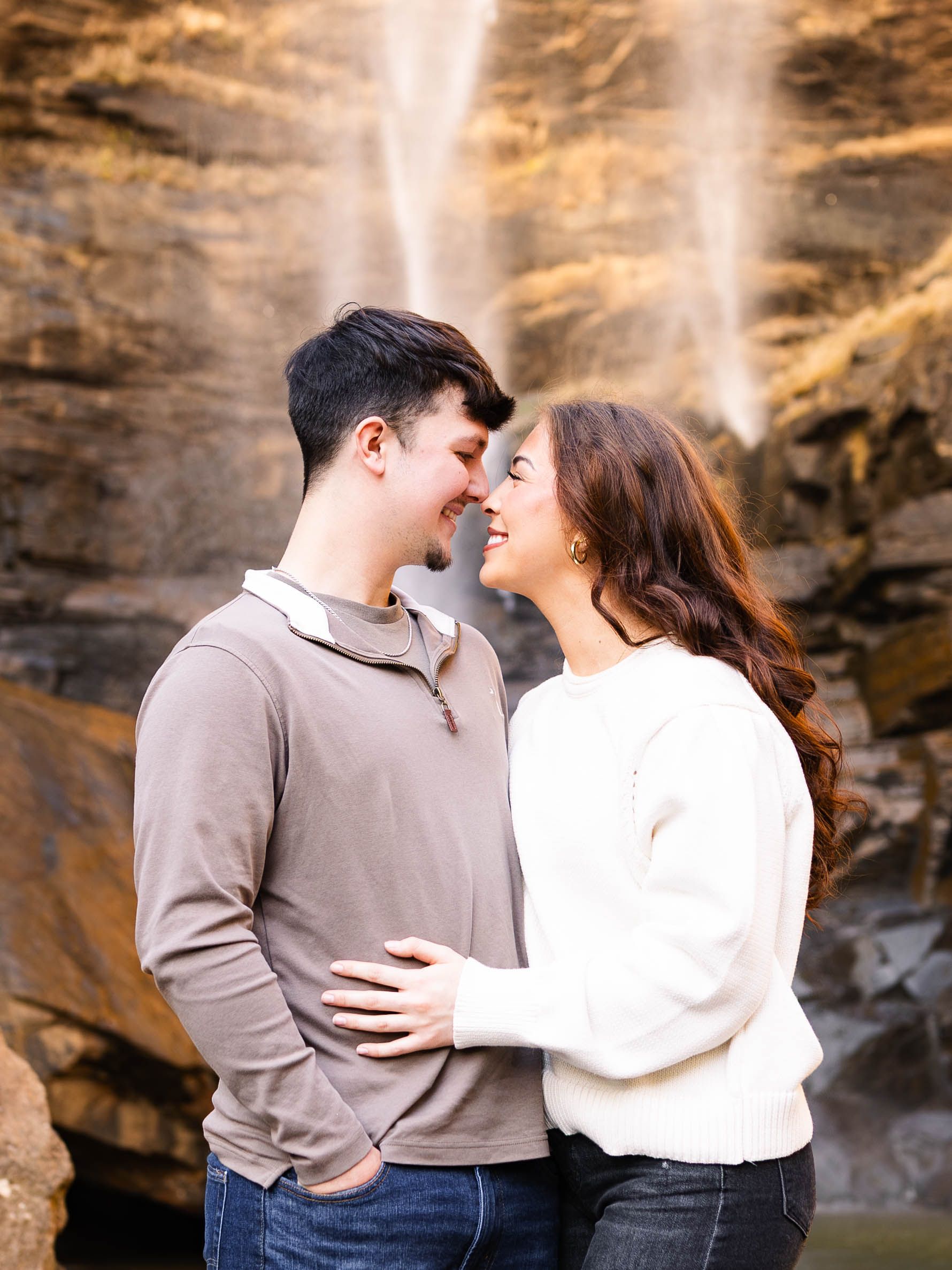 A couple shares an intimate moment against a dramatic rock wall backdrop illuminated by warm sunlight.