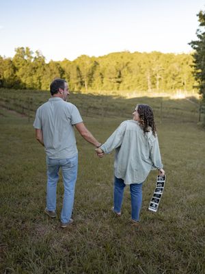 A couple holding hands while walking through a grassy field at sunset with an ultrasound photo in hand.