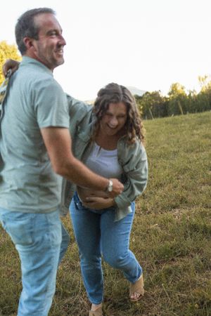 A couple laughs together while enjoying a playful moment outdoors on a grassy hillside at sunset.