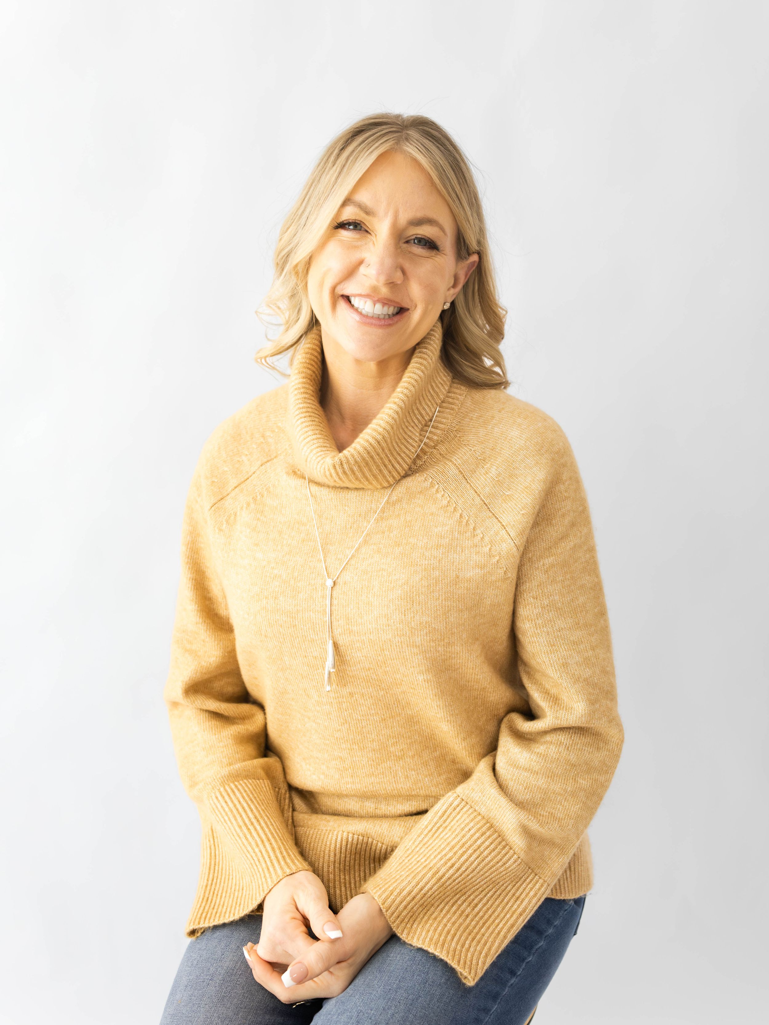 Woman wearing beige sweater smiles warmly while sitting against light background in casual lifestyle portraits.
