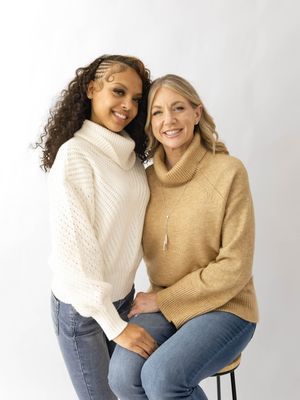 Two friends in cozy sweaters and jeans share joyful moments while sitting together against a light background.