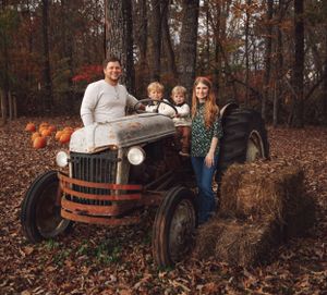 Family poses with vintage red tractor in wooded autumn setting with fallen leaves.