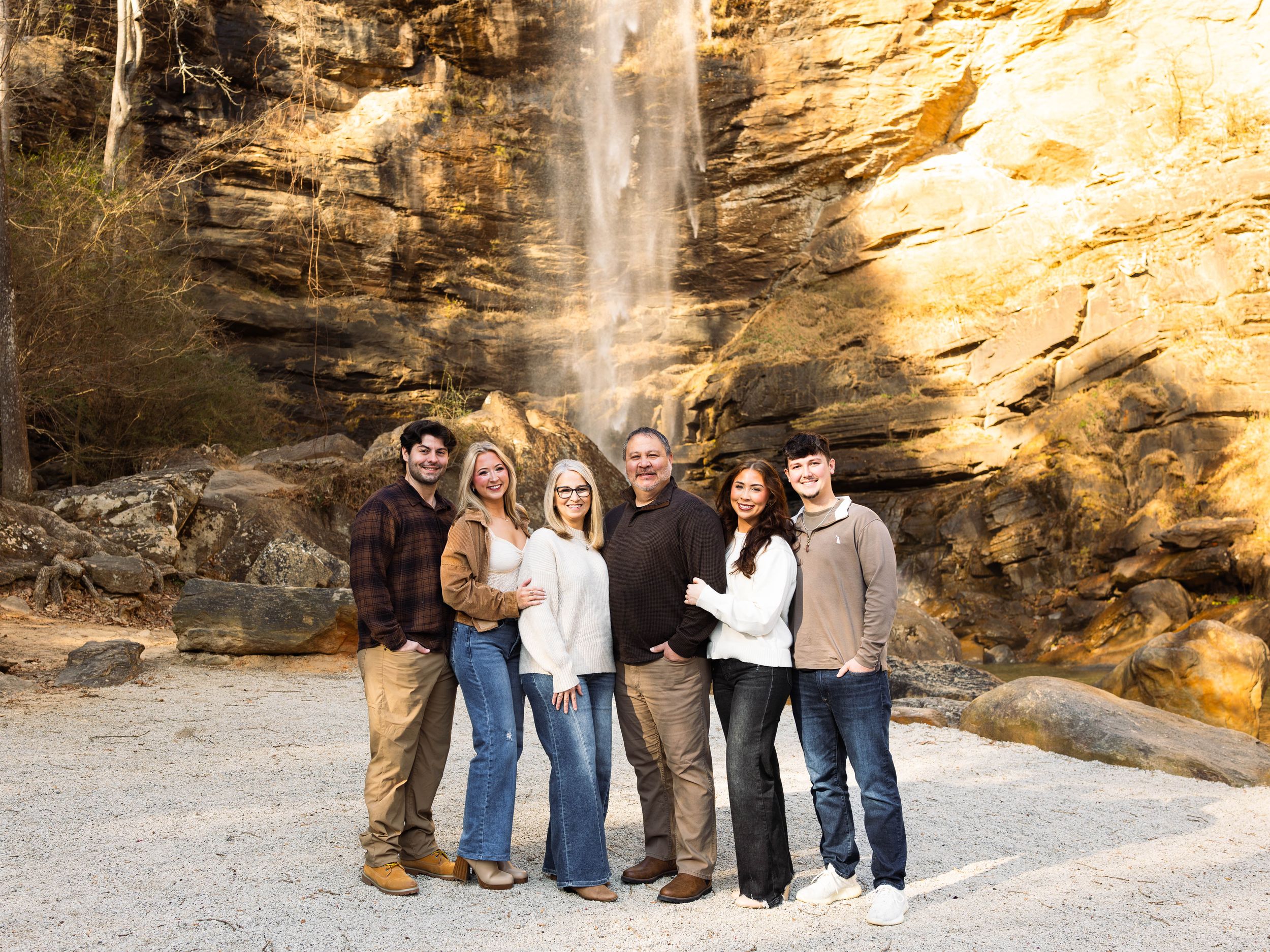 A group stands together at the base of a tall waterfall cascading down golden rocky cliffs on a sunny day.