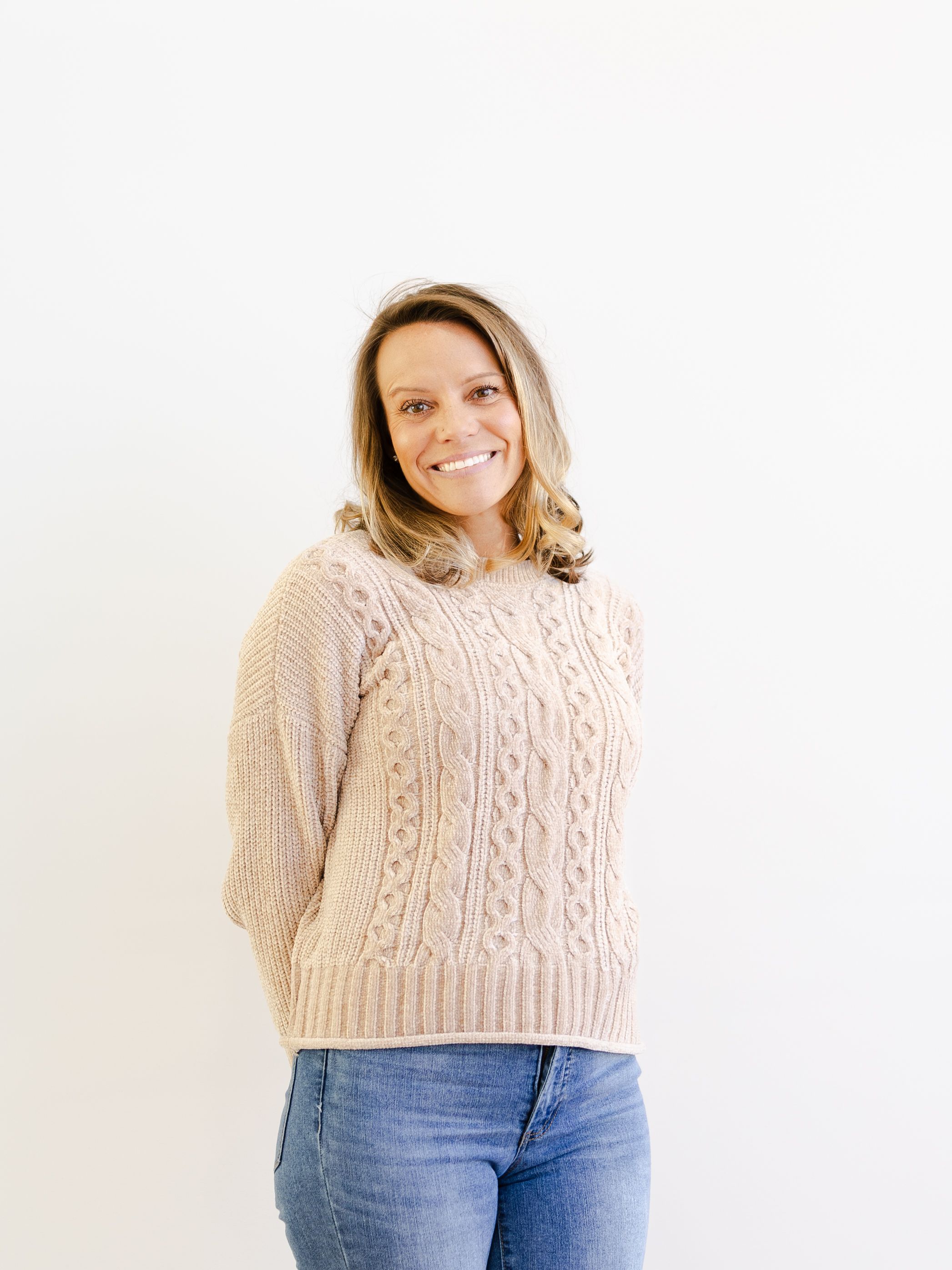 Someone wearing a cream cable knit sweater and blue jeans poses against a white background in three similar shots.