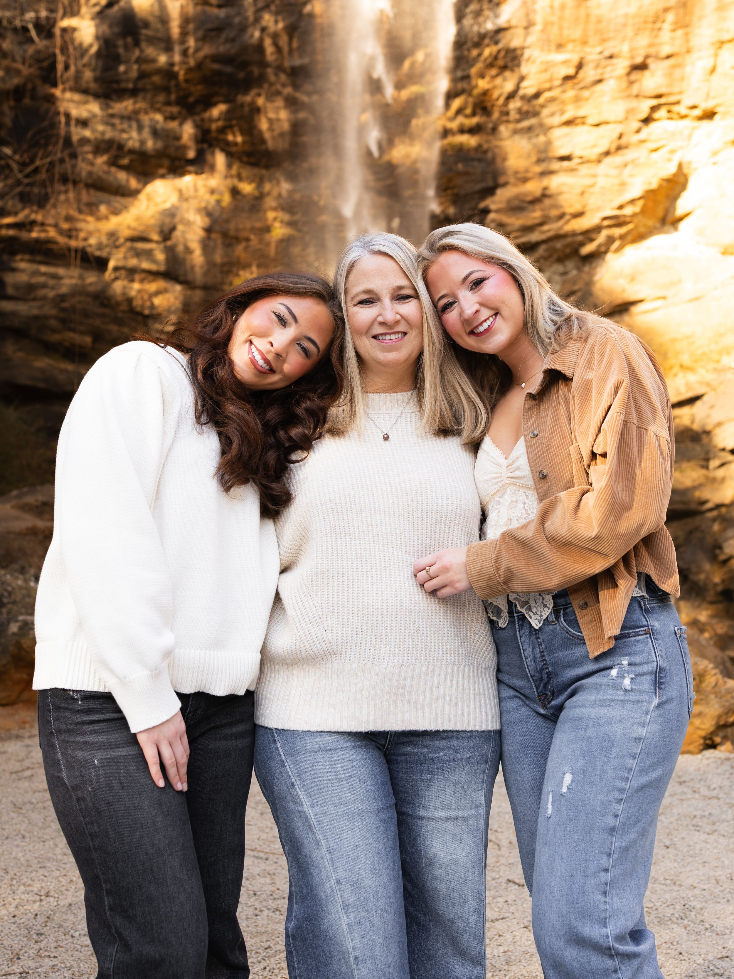 Three friends in casual outfits laugh and embrace while posing against a rocky wall backdrop.