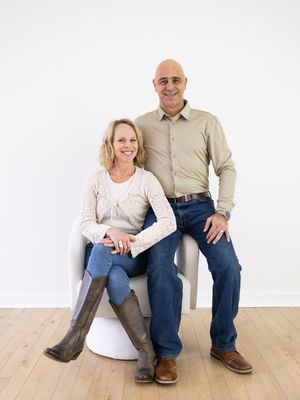 Two people sitting casually on a wooden floor wearing jeans and light colored tops against a white wall.