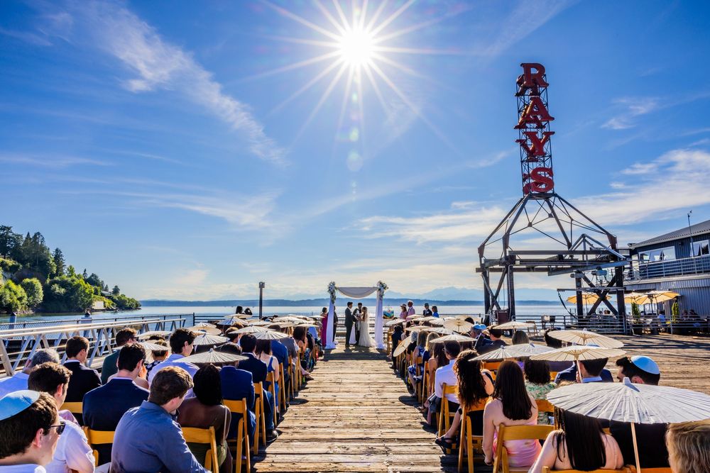 People gather at a sunny waterfront deck with blue umbrellas and a metal tower structure in Seattle's Pike Place Market.