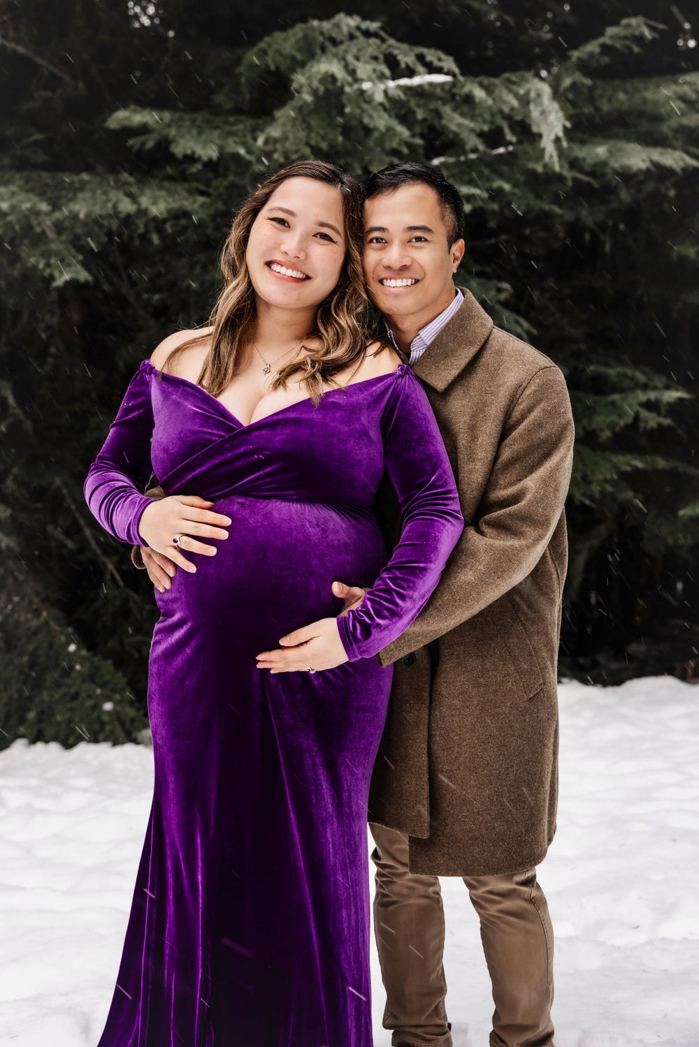 A couple poses in winter snow, with a person wearing a purple velvet maternity dress and another in a brown coat.