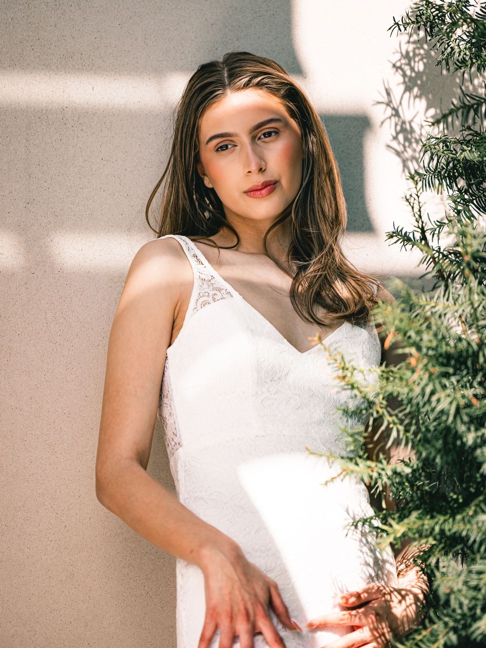 Elegant bride in white lace sleeveless wedding dress poses beside greenery in soft natural lighting.