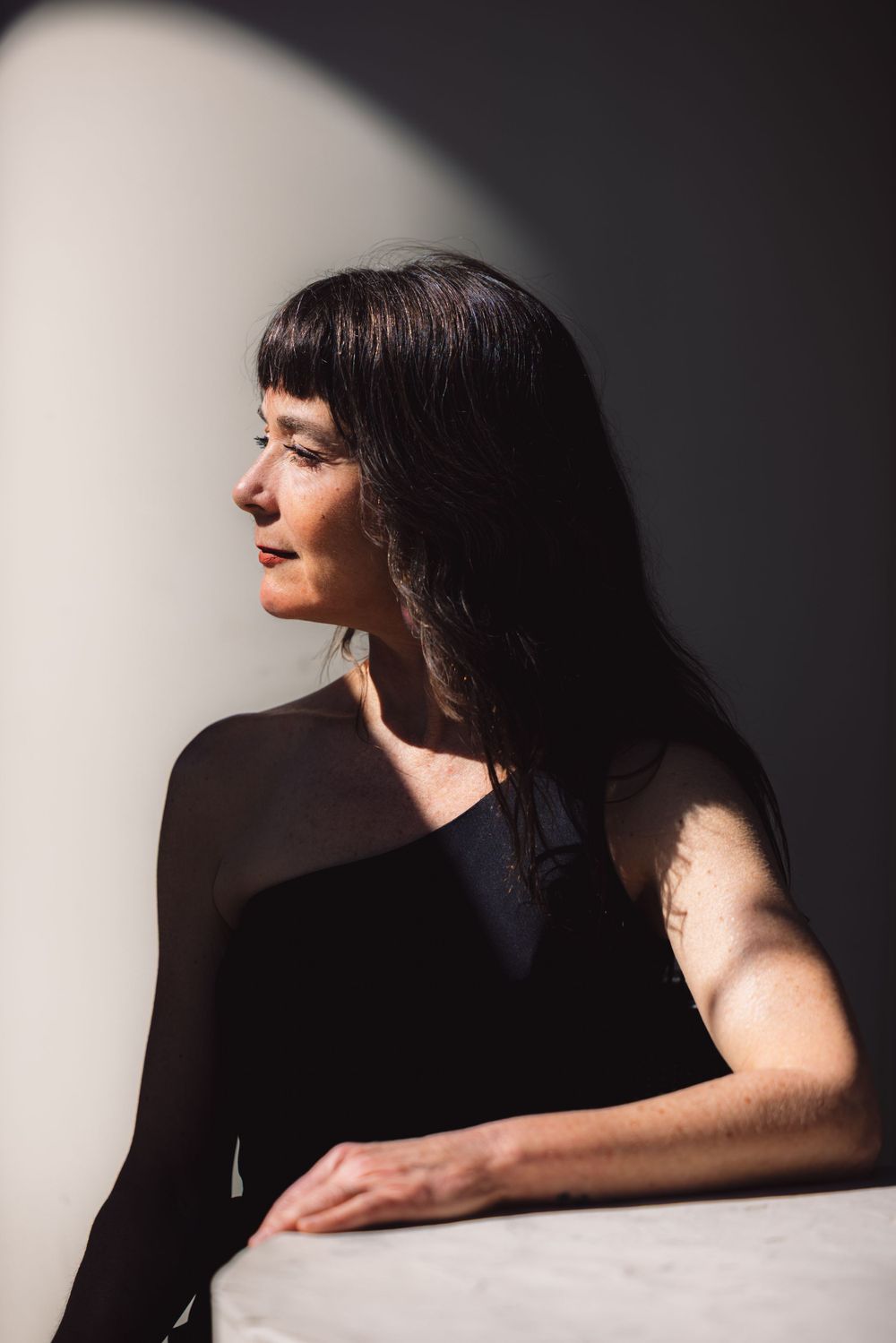 Side profile portrait of woman in black dress sitting in dramatic lighting with shadow cast on white wall.