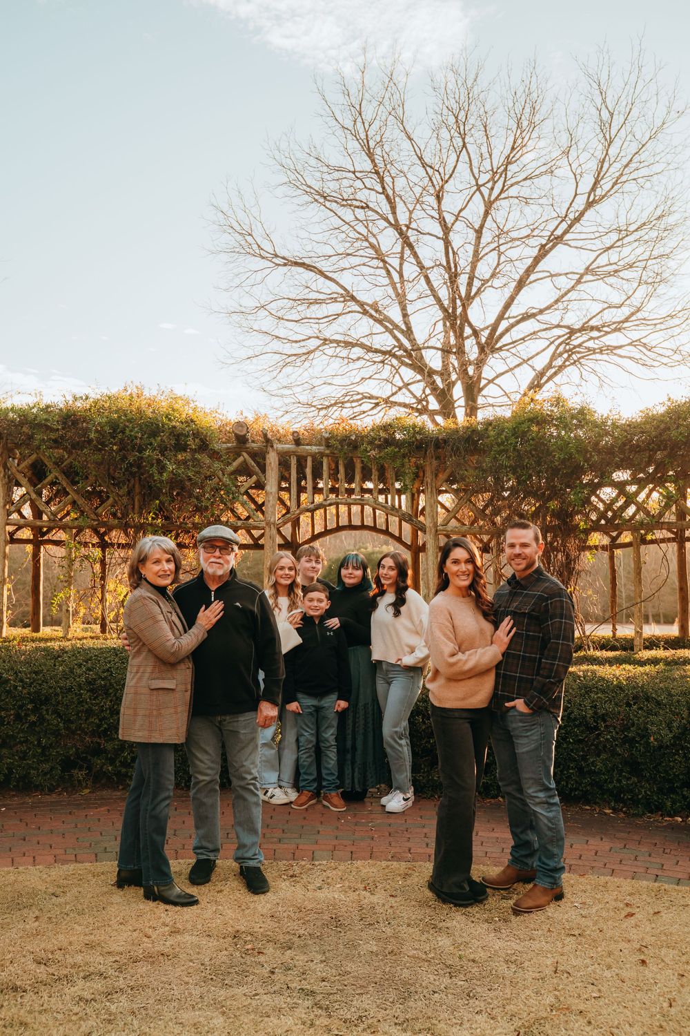 A group of people stand together in a line outdoors during golden hour against a garden backdrop with bare trees.