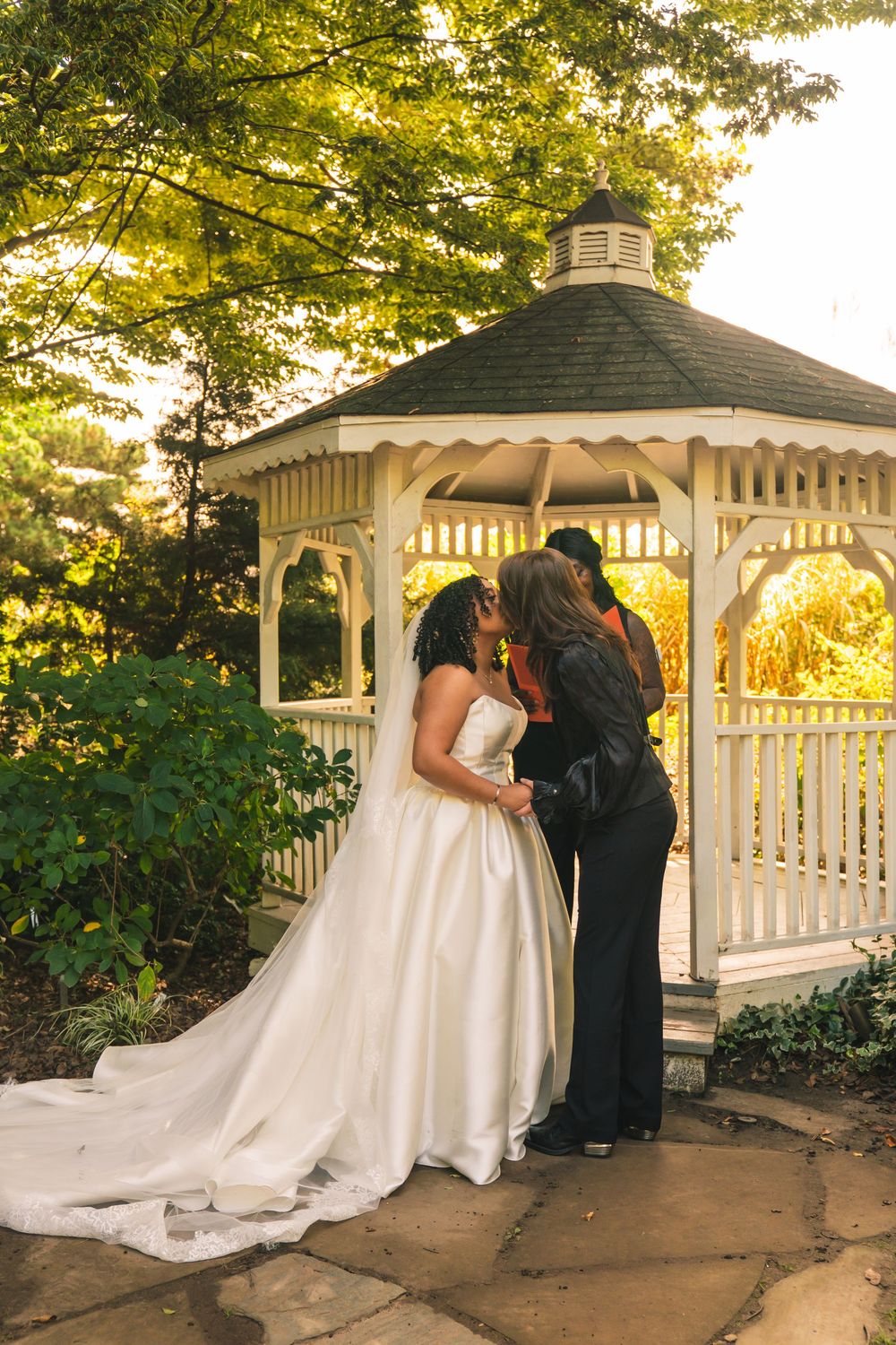 A couple shares an intimate moment in a white gazebo at sunset surrounded by lush green trees.