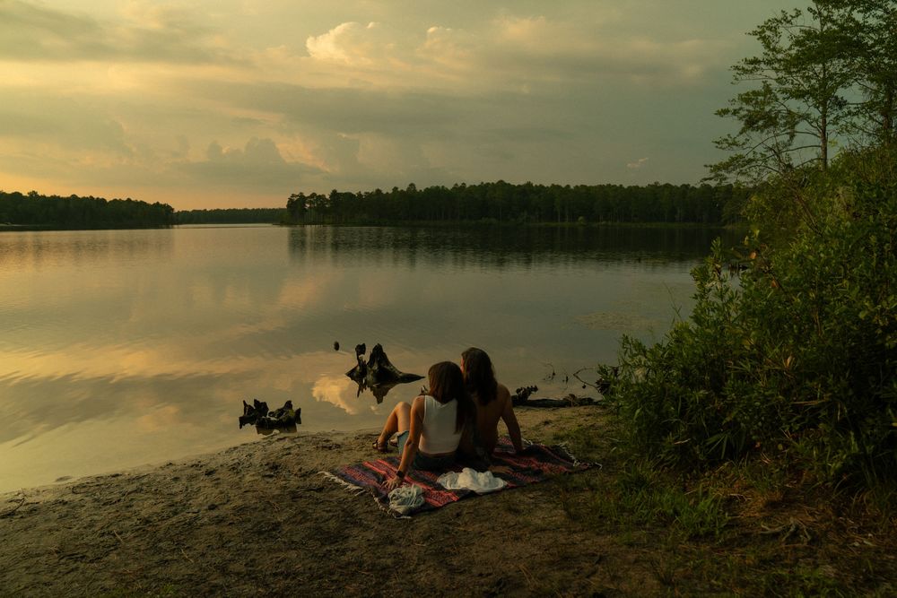 Silhouettes of two people with a dog sitting peacefully by a serene lake at sunset, surrounded by trees.