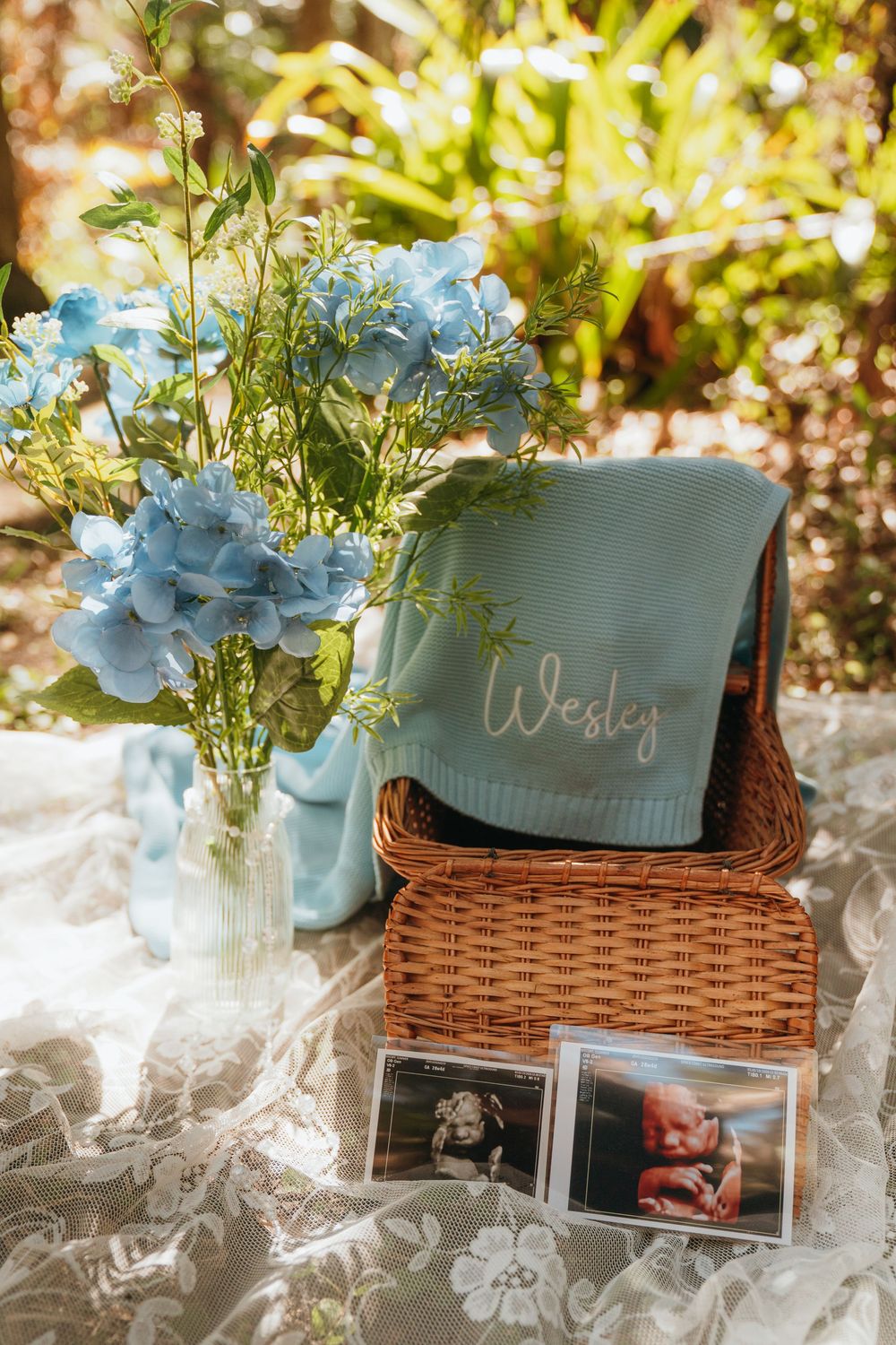 Baby announcement display with light blue flowers in vase, personalized blanket in wicker basket, and ultrasound photos on lace.