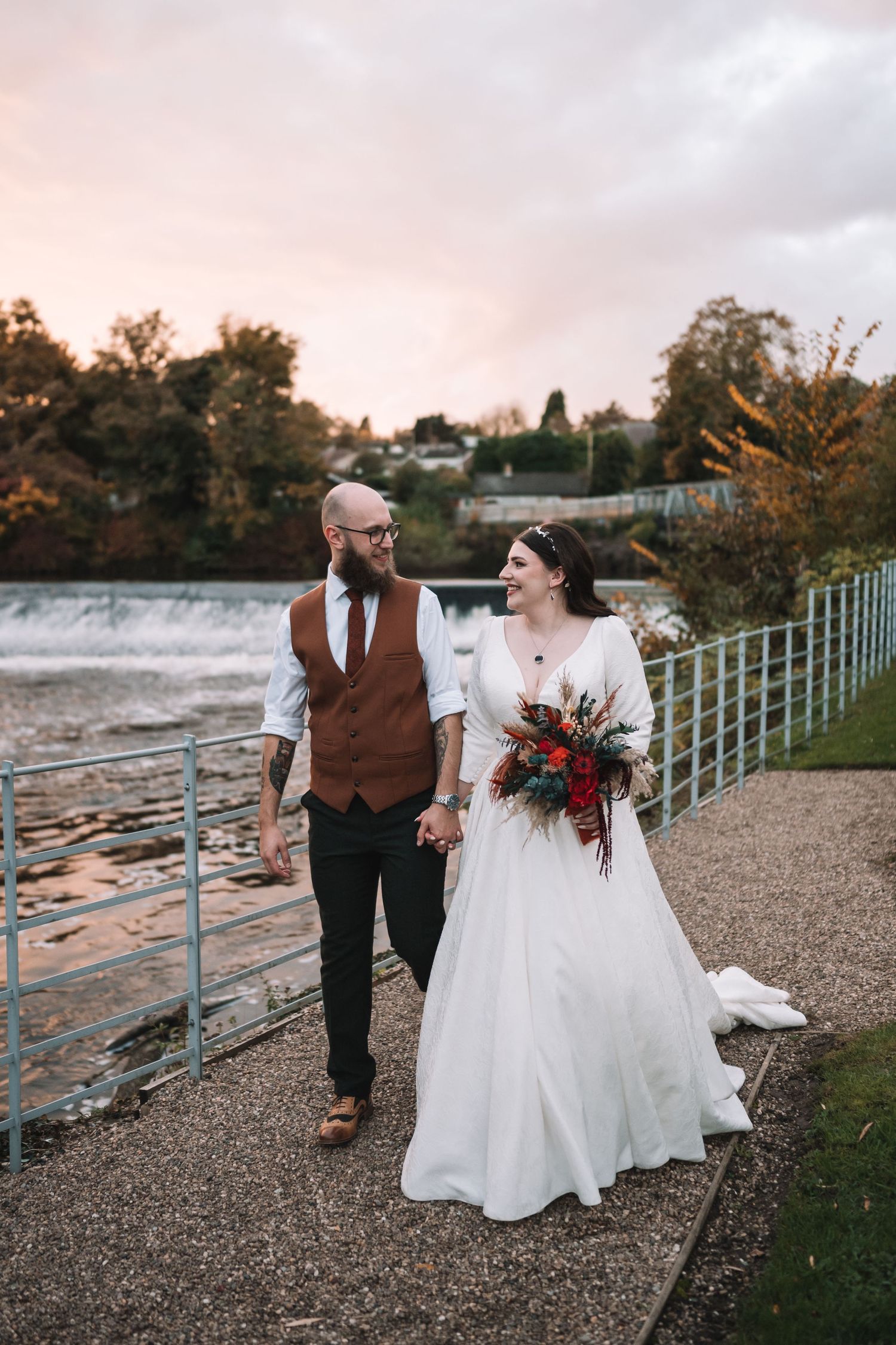 A couple walks along a waterfront path at sunset, with autumn trees and a waterfall in the background at the West Mill, Derby.