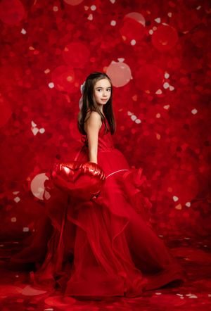 Dramatic portrait in flowing red gown with heart balloons against red bokeh background.