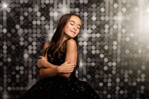 Glittering sparkly backdrop frames a person in an elegant black formal dress posing with closed eyes and crossed arms.