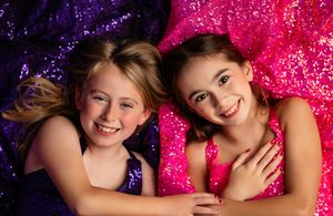 Two smiling children in sparkly purple and pink outfits pose together against a glittery backdrop.