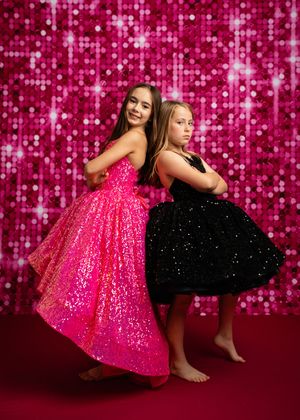 Two dancers in sparkly dresses pose against a pink sequined backdrop during a photo session.