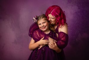 Two people share a sweet embrace wearing purple dresses against a dark purple backdrop.