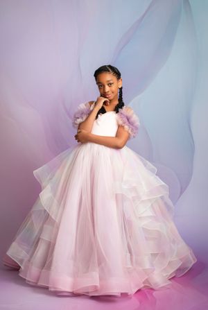 A young person poses in a voluminous white ruffled princess gown with puffy sleeves against a soft pink and purple backdrop.