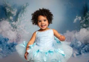 A young child wearing a light blue tulle dress smiles joyfully against a dreamy clouded backdrop.