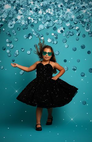 A black cocktail dress is photographed against a turquoise background with falling water droplets and sparkling effects.